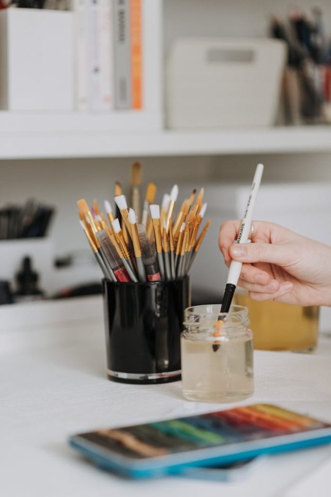 An artist's hand cleans a paintbrush in water, surrounded by more brushes and watercolor paints.